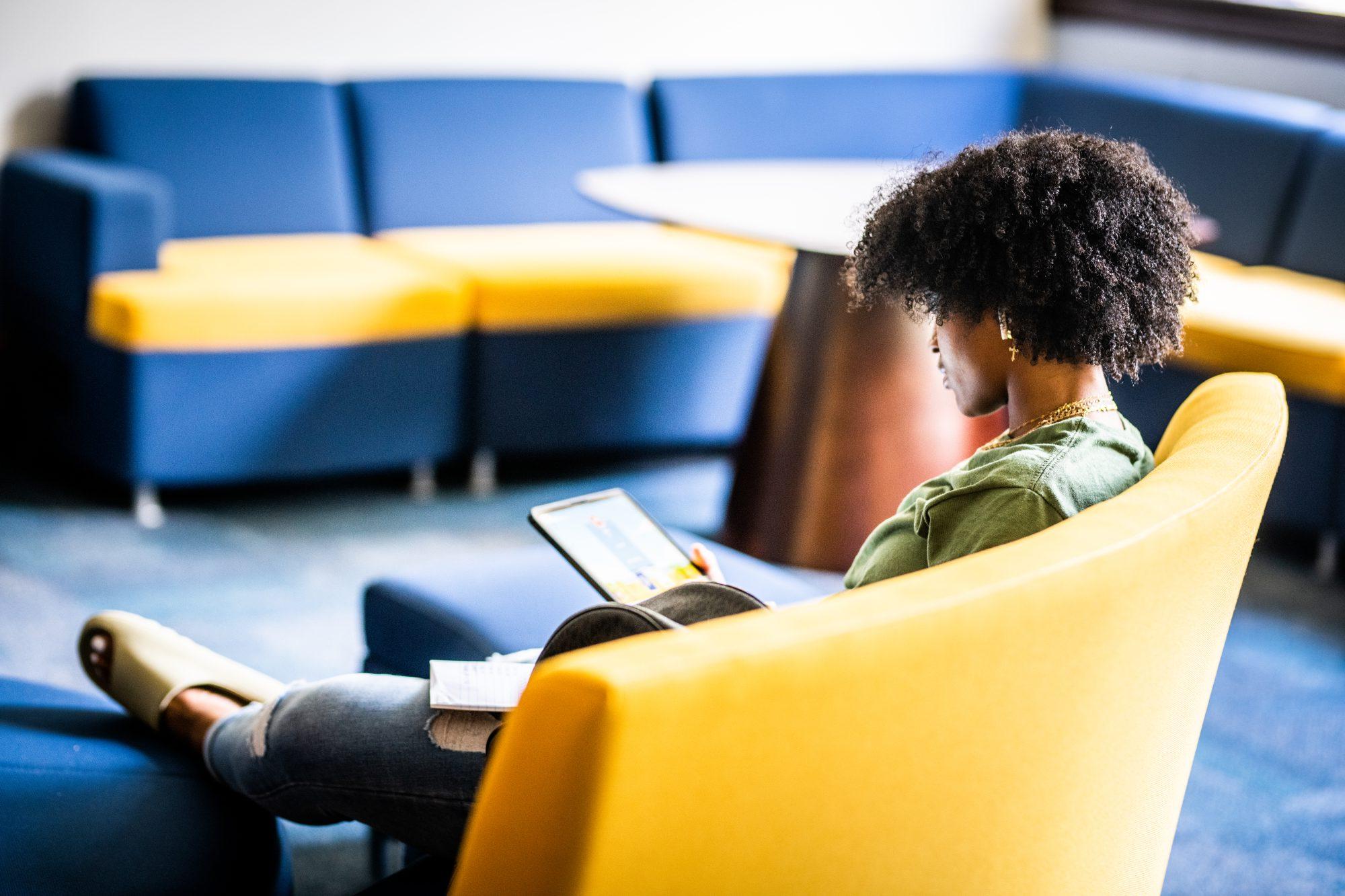 A student sits in a lounge area with a yellow and blue color scheme, focused on a tablet in their lap.